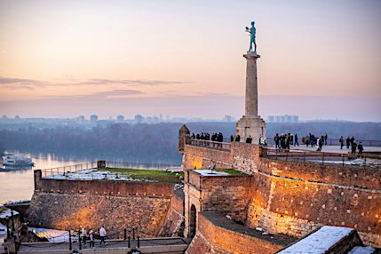Staty vid Kalemegdanfästningen med utsikt över flod och stad i solnedgång.